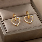 Gold huggie earrings featuring a heart drop design, accented with clear rhinestones, displayed on a beige jewelry box.