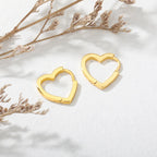 Chunky gold huggie earrings featuring a heart drop design and smooth enamel finish, set against a light, textured background.