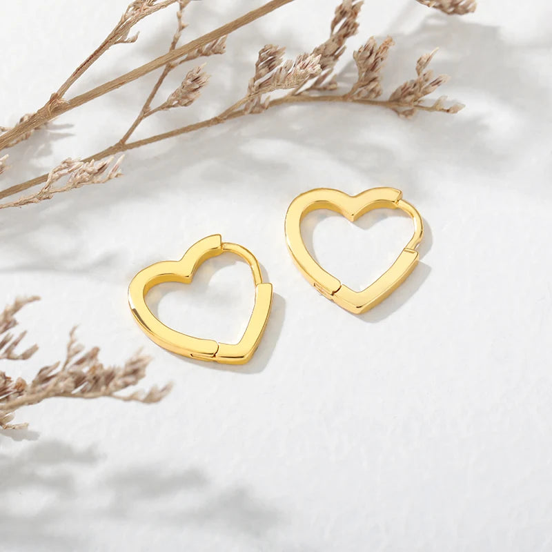 Chunky gold huggie earrings featuring a heart drop design and smooth enamel finish, set against a light, textured background.