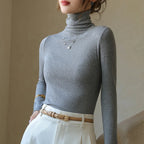 Slim-fit, gray ribbed mock-neck pullover with long sleeves, worn with high-waisted cream trousers and a belt.