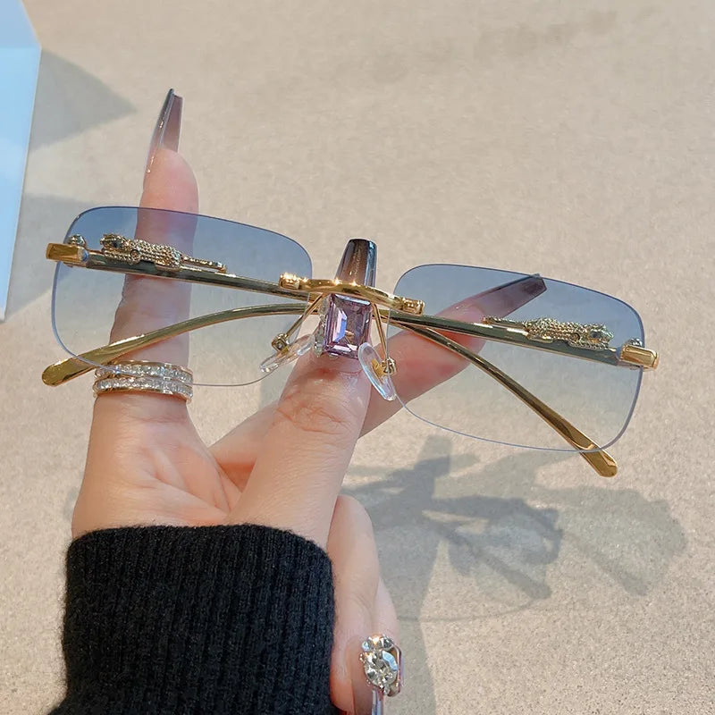Rimless Y2K rectangle sunglasses with blue gradient lenses and gold metallic accents, held between fingers.