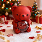 Red teddy bear holding a clear ball with a preserved rose inside, set against a festive background with holiday decorations.