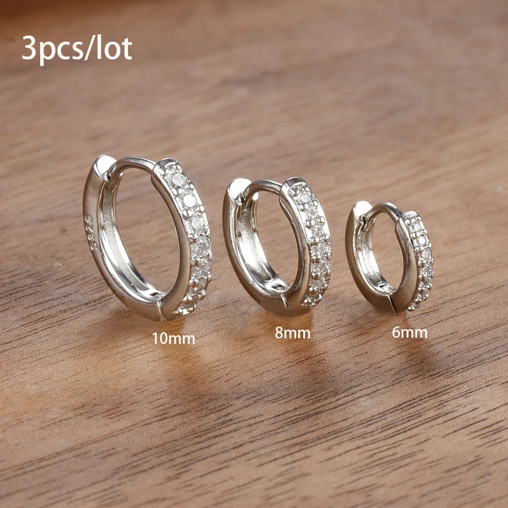 Three pairs of stainless steel hoop earrings in varying sizes, featuring sparkling stones, displayed on a wooden surface.