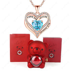 Luxury heart necklace set featuring a rose gold heart pendant with blue stone, alongside a red bear and gift box.