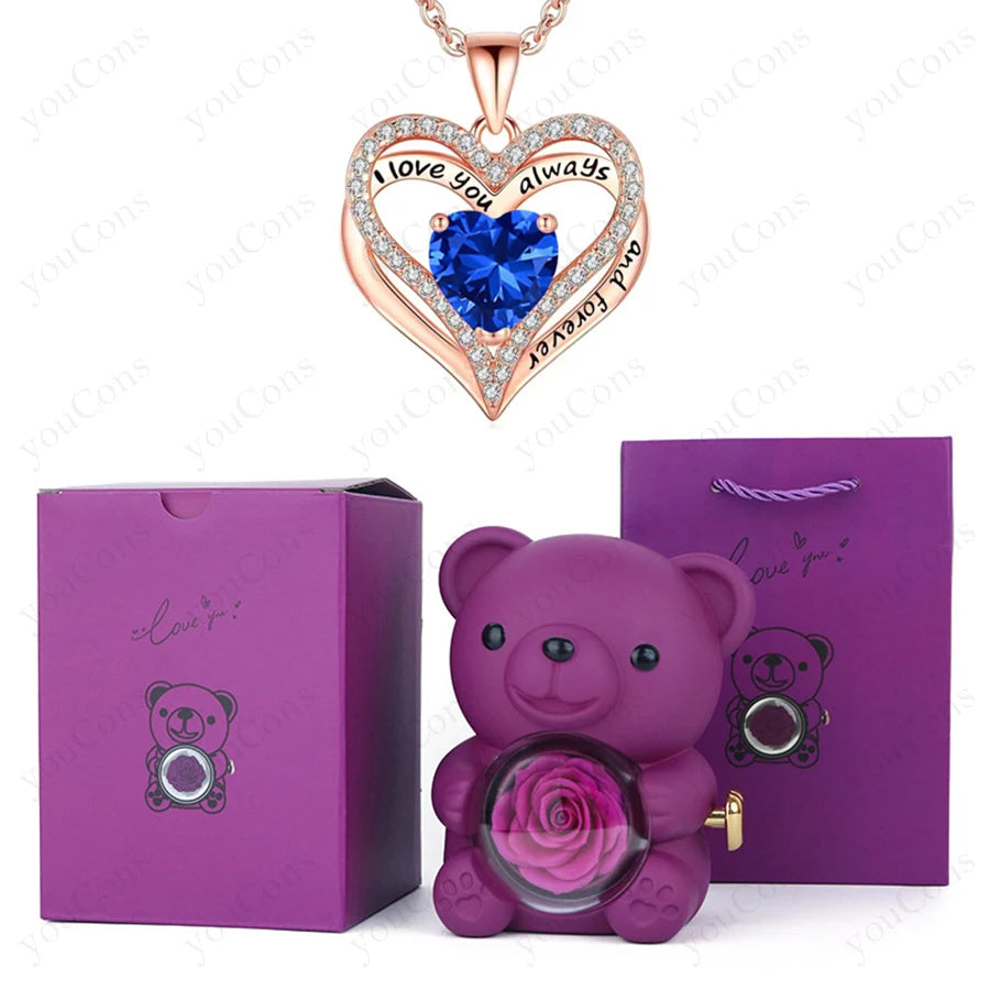 Heart-shaped necklace featuring a blue gemstone, surrounded by crystals, alongside a purple teddy bear with a preserved rose.