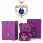 Heart-shaped necklace featuring a blue gemstone, surrounded by crystals, alongside a purple teddy bear with a preserved rose.