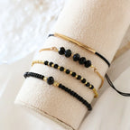 Four-piece stackable bracelet set featuring gold-tone beads and black onyx glass stones, displayed on a neutral cushion.