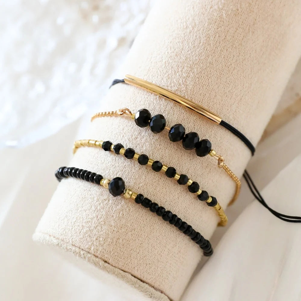 Four-piece stackable bracelet set featuring gold-tone beads and black onyx glass stones, displayed on a neutral cushion.