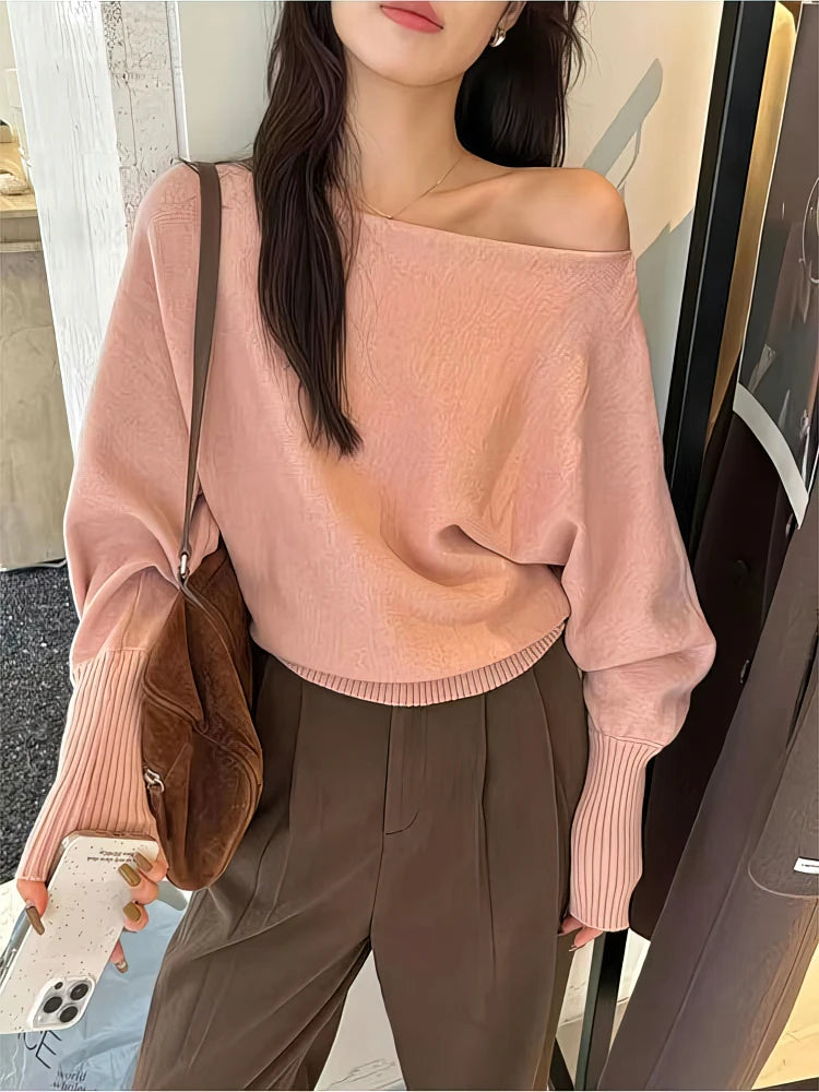Soft pink LOVORA™ Off-Shoulder Batwing Knit Pullover with relaxed long sleeves, worn off one shoulder, paired with brown pants.