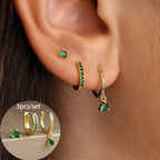 Three-piece earring set featuring gold-toned geometric designs, including a stud, a hoop with green gems, and a dangle.