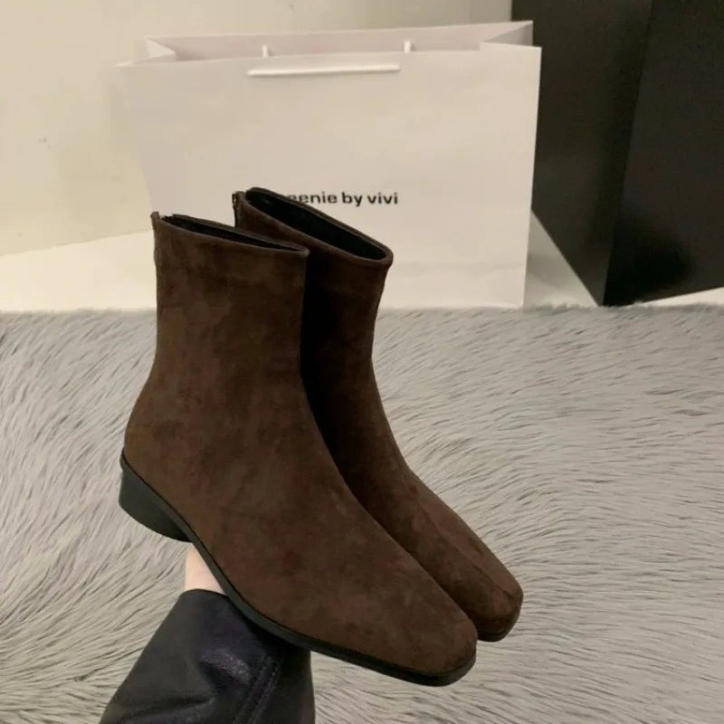 Brown suede ankle boots with a low-profile stacked block heel and back zipper, held against a soft surface.