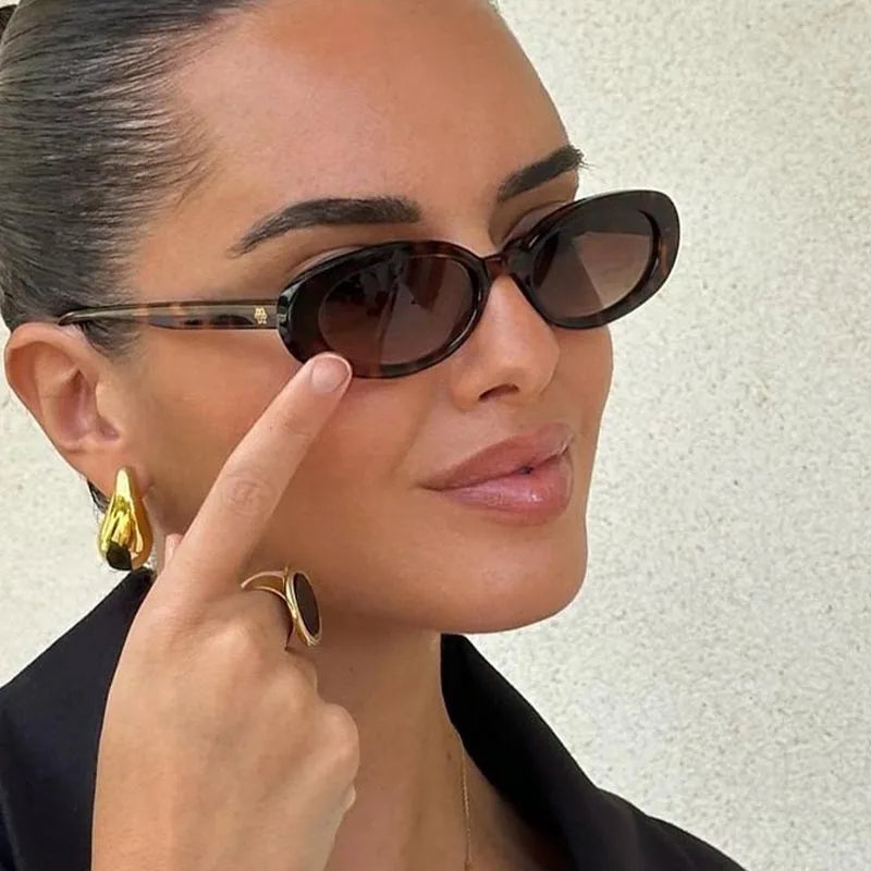 Vintage oval sunglasses with a tortoiseshell pattern, shown on a model pointing at them, paired with gold jewelry.