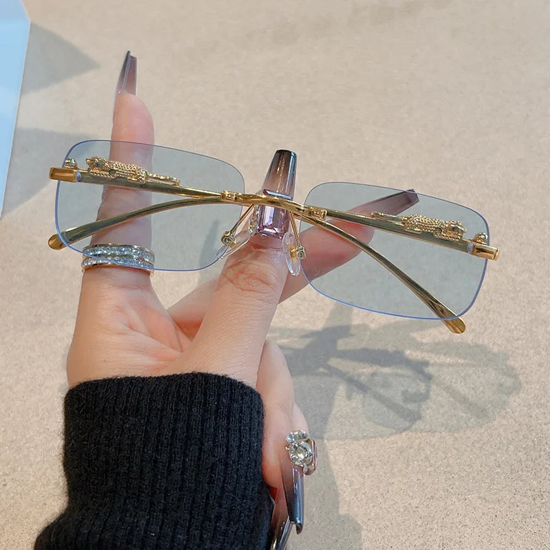 Rimless Y2K rectangle sunglasses with gradient blue lenses, gold metallic temples, held in a hand with rings.