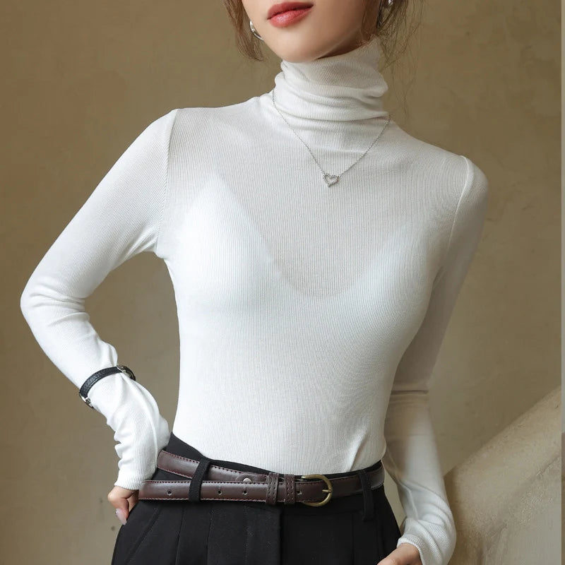 Slim-fit white ribbed mock-neck pullover with long sleeves, worn with a black belt and high-waisted pants.