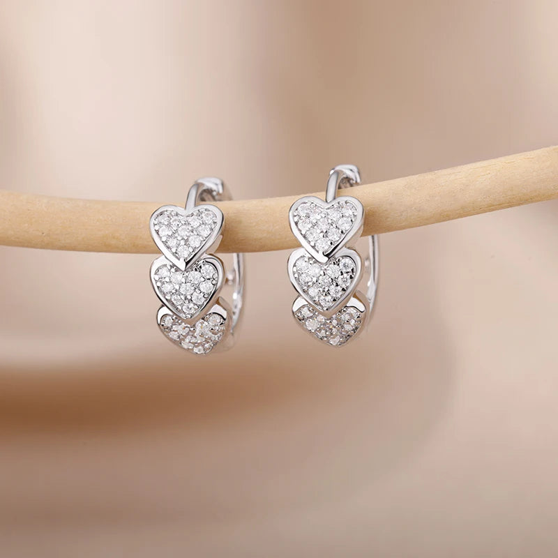 Chunky huggie hoop earrings featuring three stacked heart designs, adorned with shimmering clear stones, mounted on a soft background.
