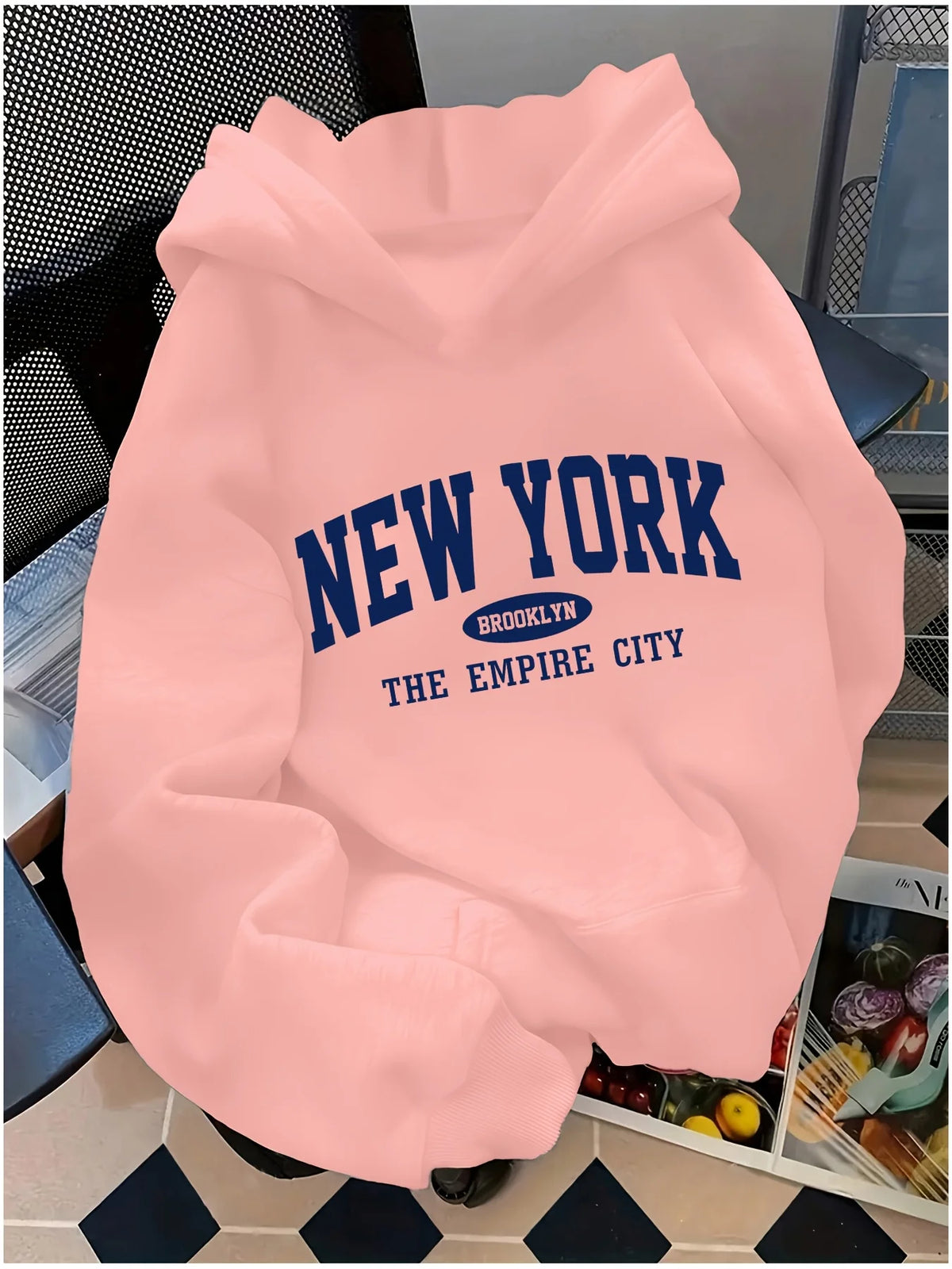 Pink oversized hoodie featuring a navy "NEW YORK" graphic, spacious hood, and kangaroo pocket on a patterned surface.