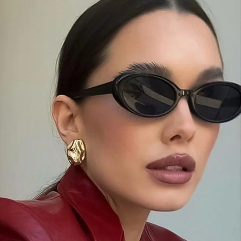 Vintage oval sunglasses with a sleek black frame, worn by a model in a crimson jacket and bold gold earrings.