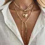 Layered gold necklace featuring beaded chains with a large heart pendant, worn above a white button-up shirt.