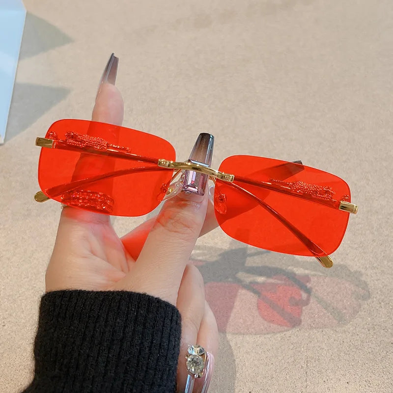 Rimless Y2K rectangle sunglasses with gradient red lenses and gold metallic hinges, held by a hand with long nails.