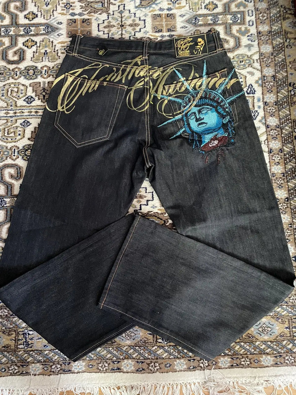 Baggy distressed denim jeans in dark wash with blue embroidered Statue of Liberty and gold script design on the back.