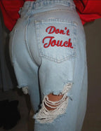 Light blue distressed denim jeans with red embroidered "Don't Touch" detail, oversized baggy fit, and frayed knee rips.