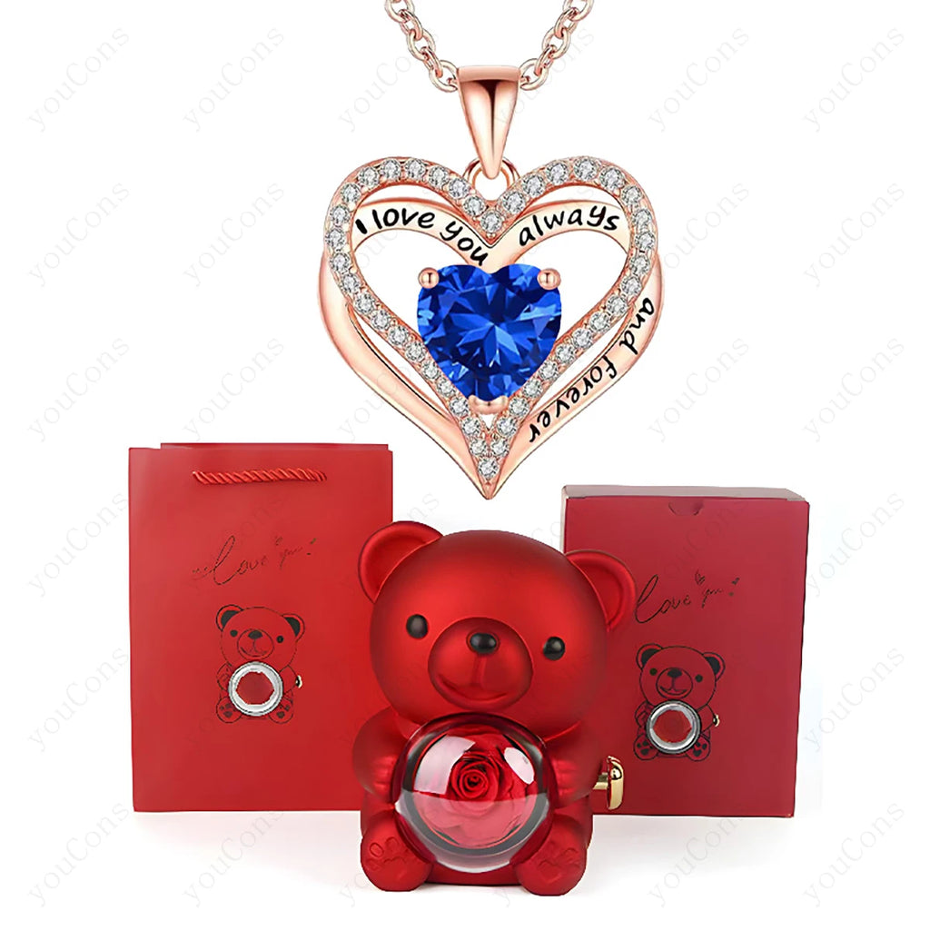 Rose gold heart necklace with blue gemstone, surrounded by sparkling crystals. Includes a red bear figurine and gift boxes.
