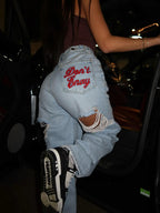 Light blue baggy distressed denim jeans with "Don't Envy" embroidery on the back pocket, styled with black sneakers.