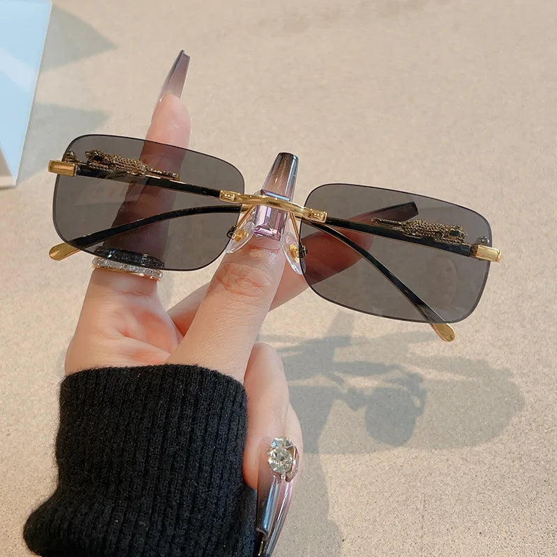 Rimless rectangle sunglasses with gradient black lenses, metallic hinges, and slender temples, held by a hand adorned with rings.
