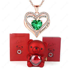 Red heart pendant necklace featuring a green gem, surrounded by crystals, with a red teddy bear and gift boxes.
