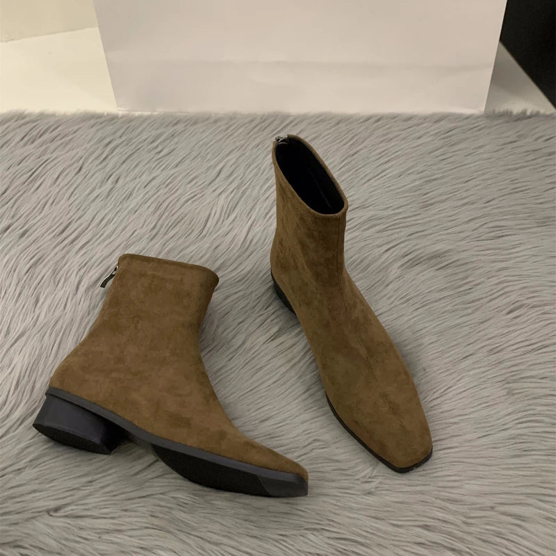 Brown suede ankle boots with a pointed toe, back zipper closure, and low stacked heel, displayed on a soft surface.