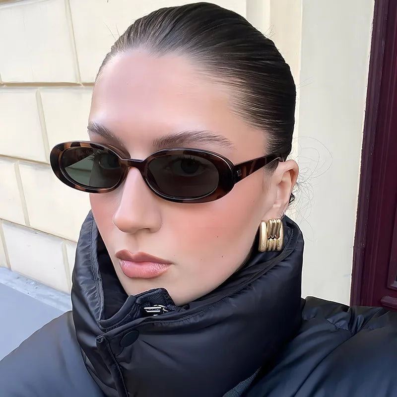 Oval minimalist sunglasses in a tortoiseshell pattern, worn by a model with a high collar black puffer jacket.