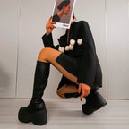 Chunky black platform boots with a curved block heel, paired with a black outfit featuring floral accents. Model holds a magazine.