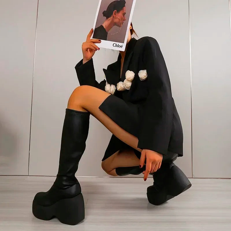 Chunky black platform boots with a curved block heel, paired with a black outfit featuring floral accents. Model holds a magazine.