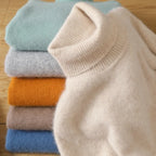 Light beige turtleneck sweater made of soft merino wool and cashmere, displayed on top of stacked colorful sweaters.