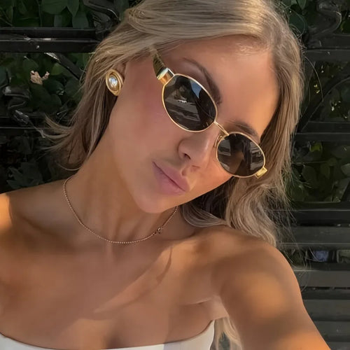 Geometric hexagonal sunglasses with a gold metal frame and dark lenses, worn by a model with long hair.