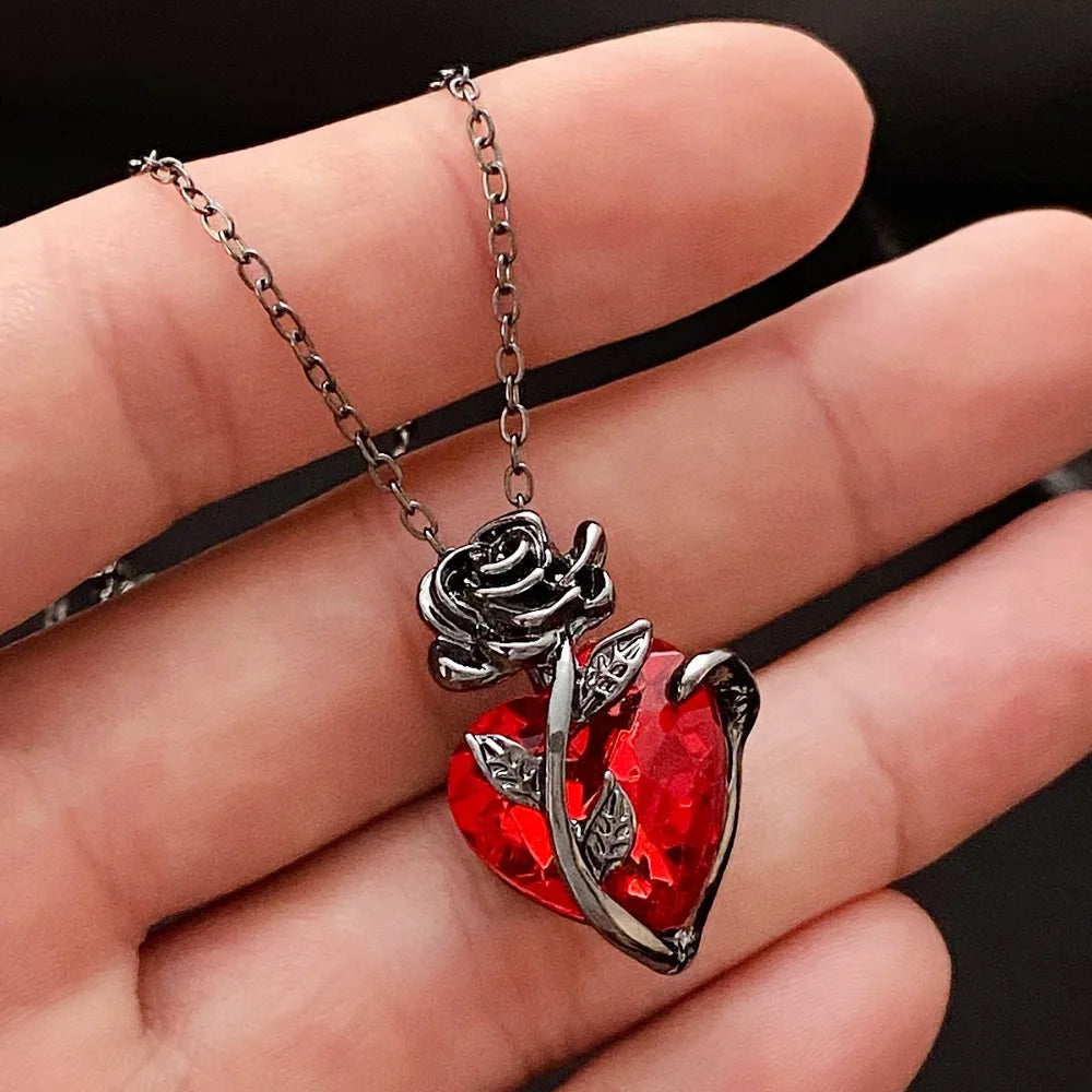 A hand holding a Gothic rose and crimson anatomical heart pendant necklace on a dark chain, featuring intricate floral metalwork.