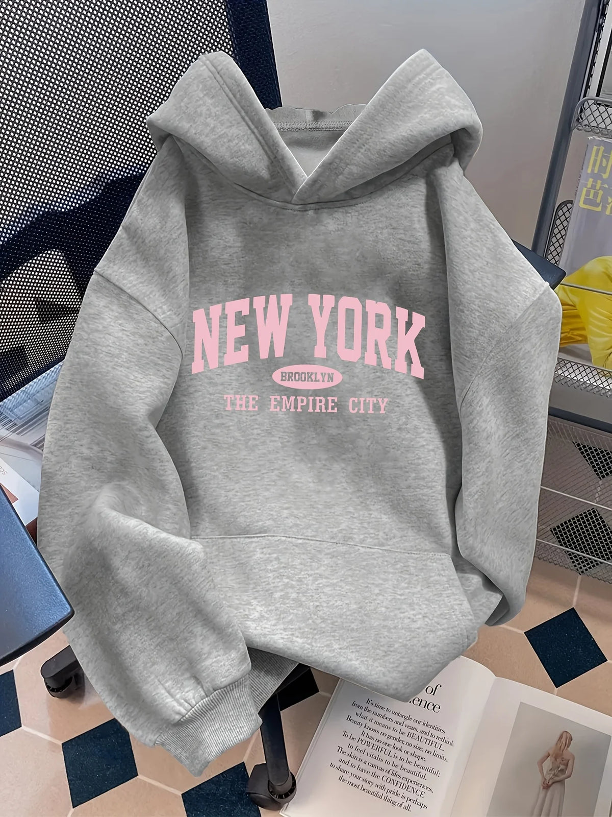 Gray oversized hoodie featuring a pink "NEW YORK" graphic. Includes a spacious hood and ribbed cuffs, styled in a casual setting.