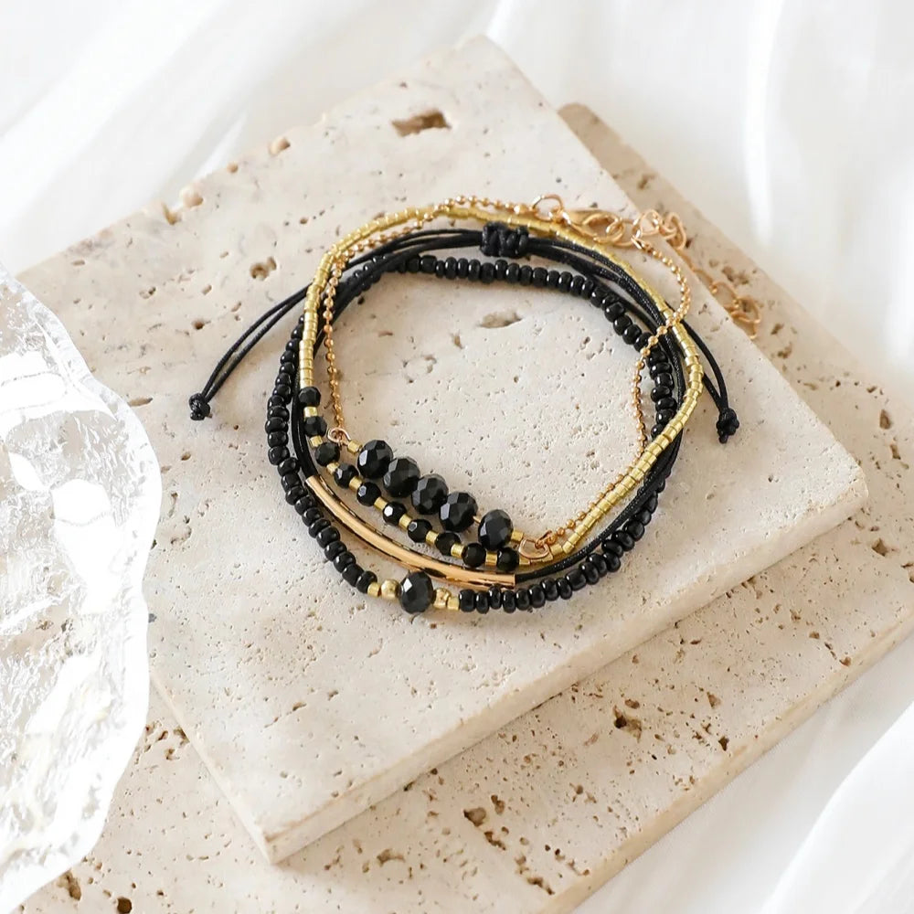 Stackable bracelet set featuring four pieces with black onyx glass stones and metallic gold-tone beads on a textured surface.