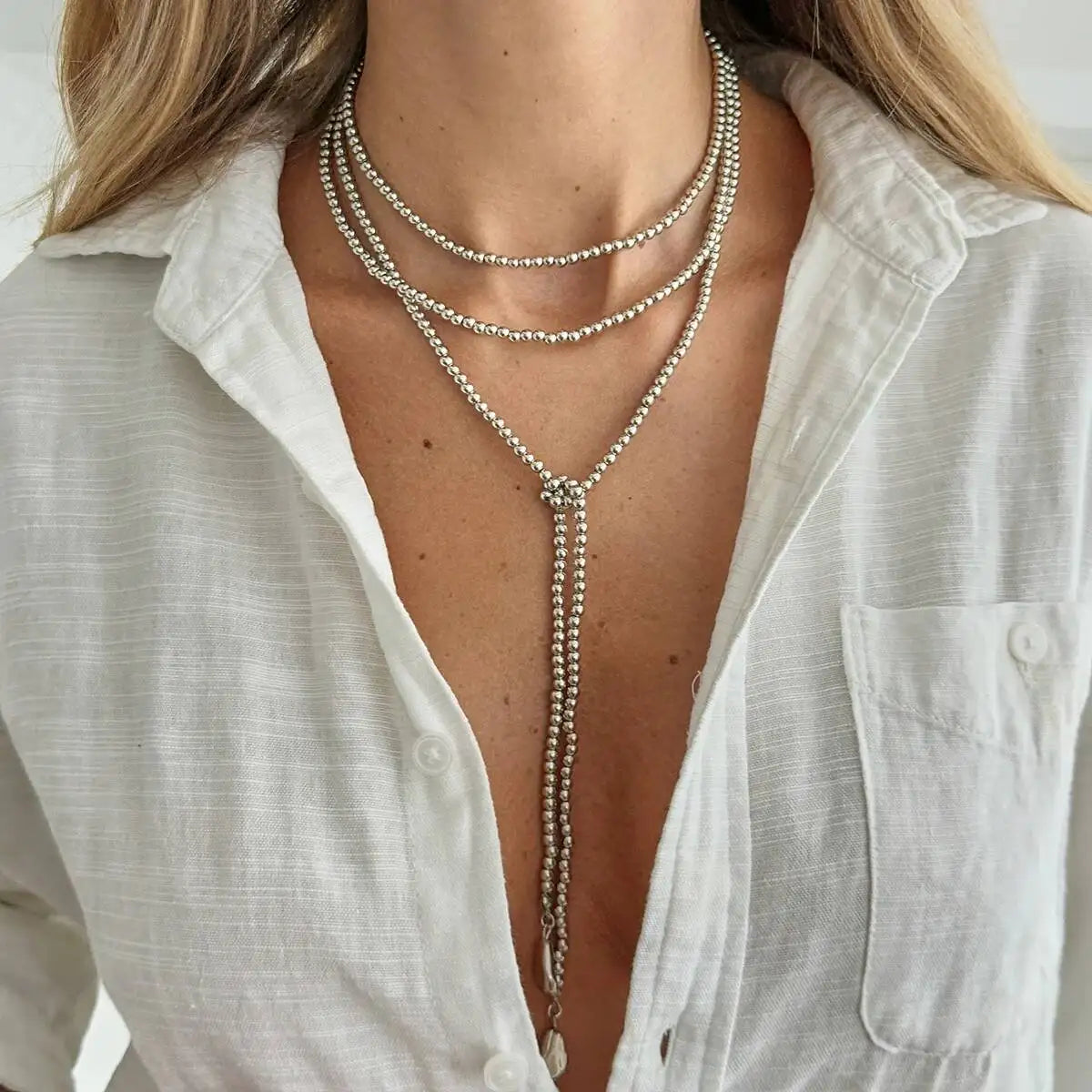 Layered necklace featuring silver beads and a heart pendant, worn over a white button-up shirt, draping elegantly.