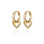 Chunky huggie earrings in gold with a heart drop design and crystal accents, set against a reflective surface.