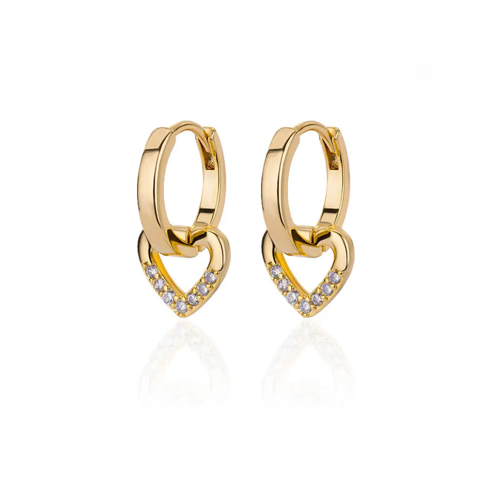 Chunky huggie earrings in gold with a heart drop design and crystal accents, set against a reflective surface.