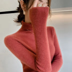 Slim-fit turtleneck sweater in rich rose color, featuring ribbed texture and high collar, styled with hands partially covering face.