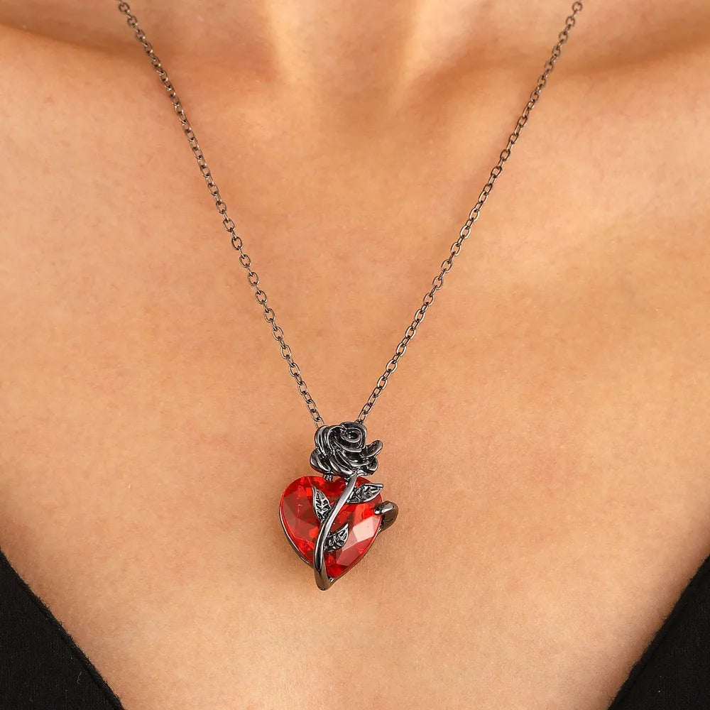Crimson anatomical heart pendant wrapped in dark rose metalwork, suspended on a delicate chain, worn at the collarbone.