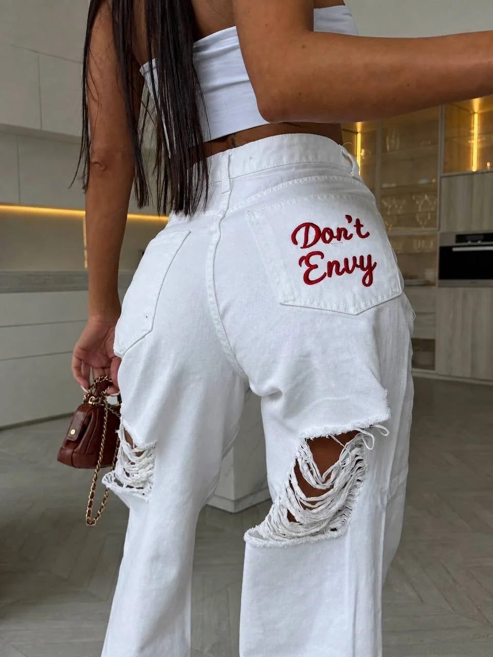 Baggy white denim jeans with distressed rips and red embroidered text "Don't Envy" on the back pocket, styled with a cropped white top.