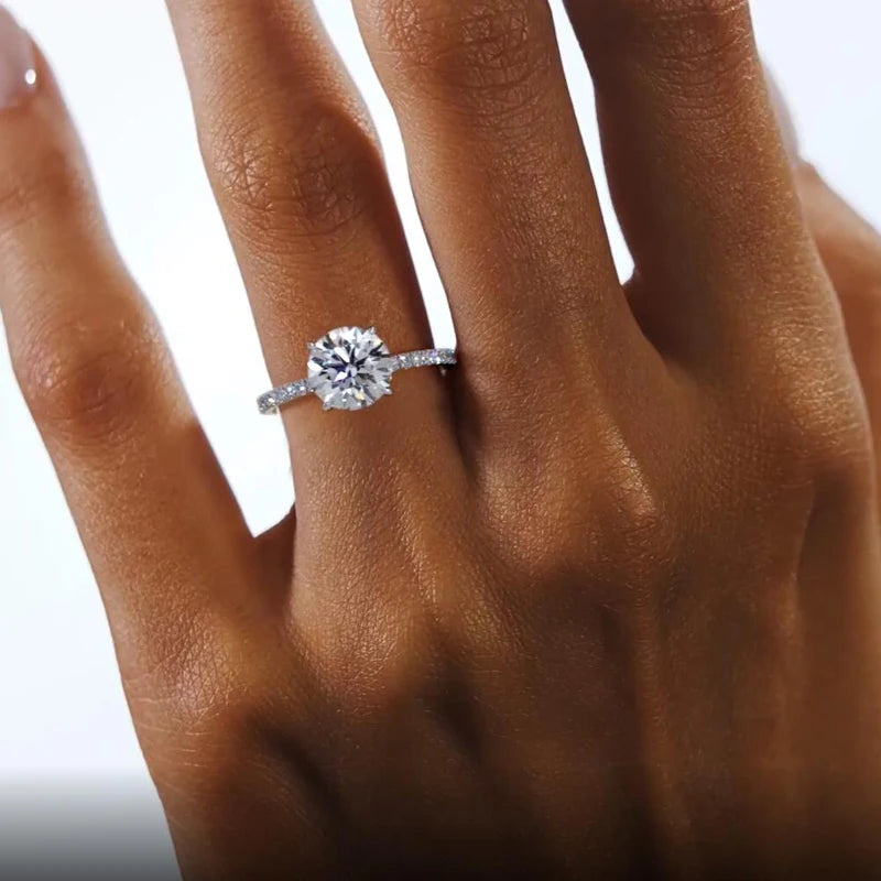 A hand displaying a 6-prong moissanite solitaire ring with a brilliant round-cut stone set on a silver band.