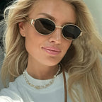 Hexagonal metal sunglasses with dark lenses worn by a model with long blonde hair, featuring a simple white t-shirt and pearl necklace.