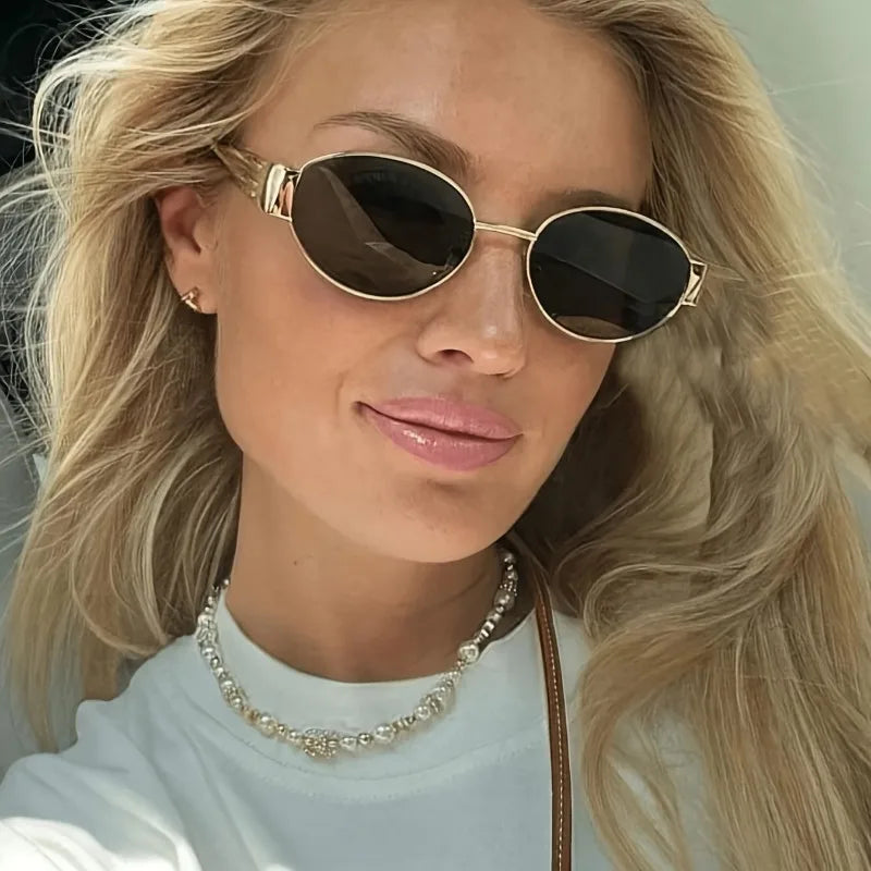 Hexagonal metal sunglasses with dark lenses worn by a model with long blonde hair, featuring a simple white t-shirt and pearl necklace.