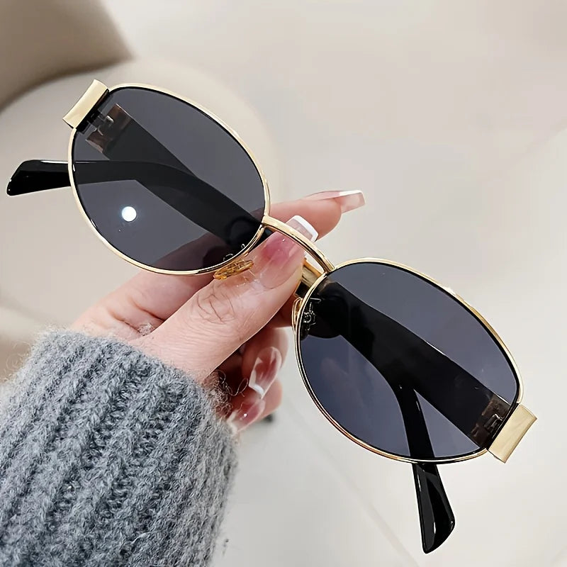 Geometric hexagonal sunglasses with a gold metal frame and black lenses, held in a hand with a gray sweater.