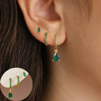 Gold-tone earring set featuring a small emerald-cut stud, a green gem-accented hoop, and a teardrop design, worn in multiple piercings.